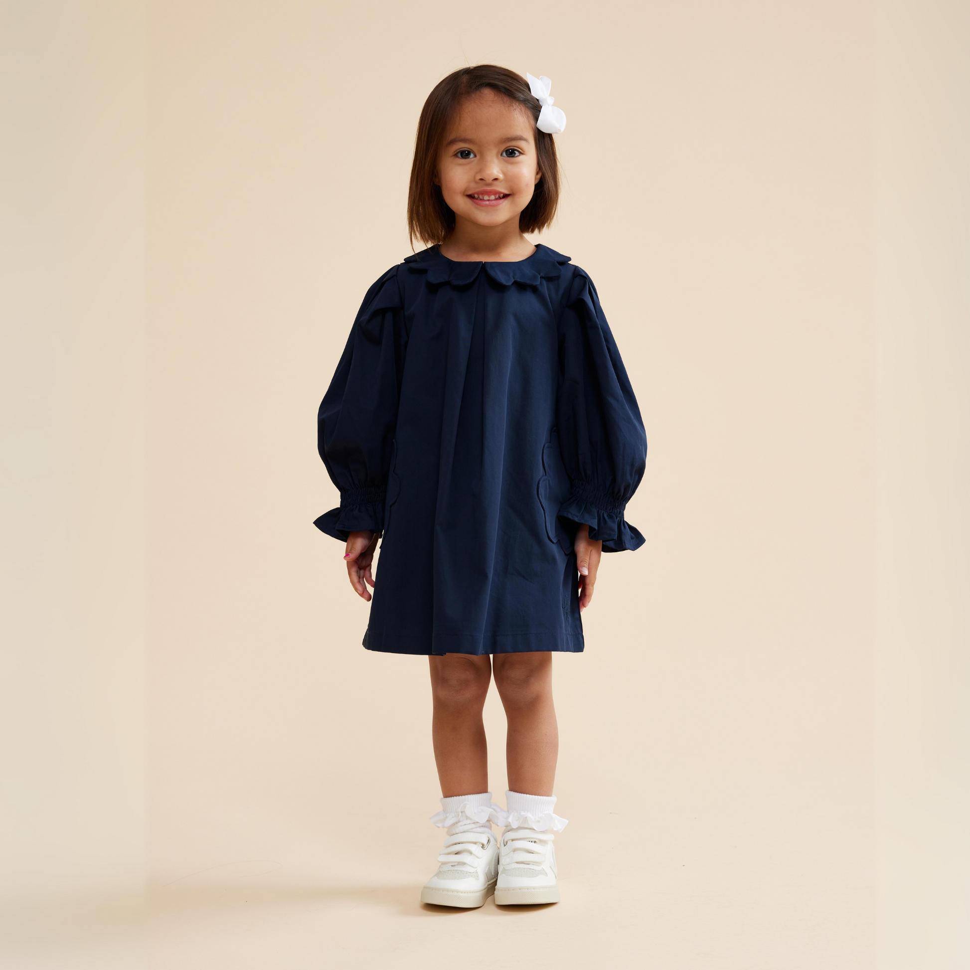 A child standing, wearing a navy blue scallop detail smock dress with short sleeves and a collar, paired with white shoes.