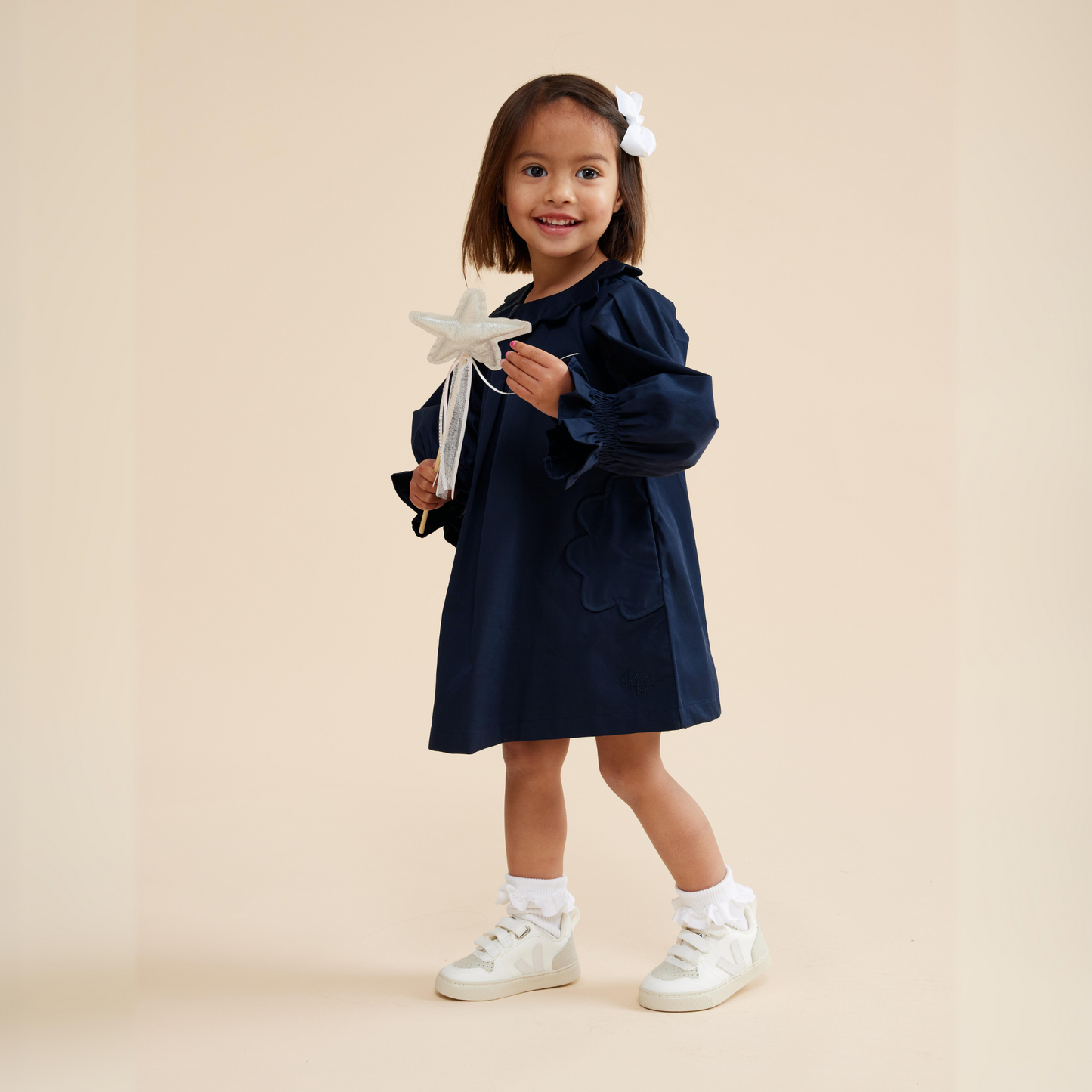 Girl wearing navy scallop detail smock dress styled with frill sock, white sneaker, white bow hair clip and fairy wand