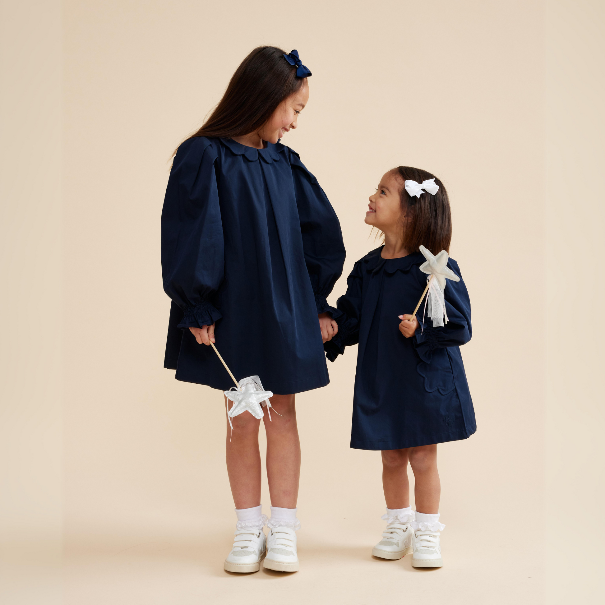 Little girls wearing navy scallop detail smock dress, showing long puff sleeves, soft cotton, paired with frill socks and white sneakers