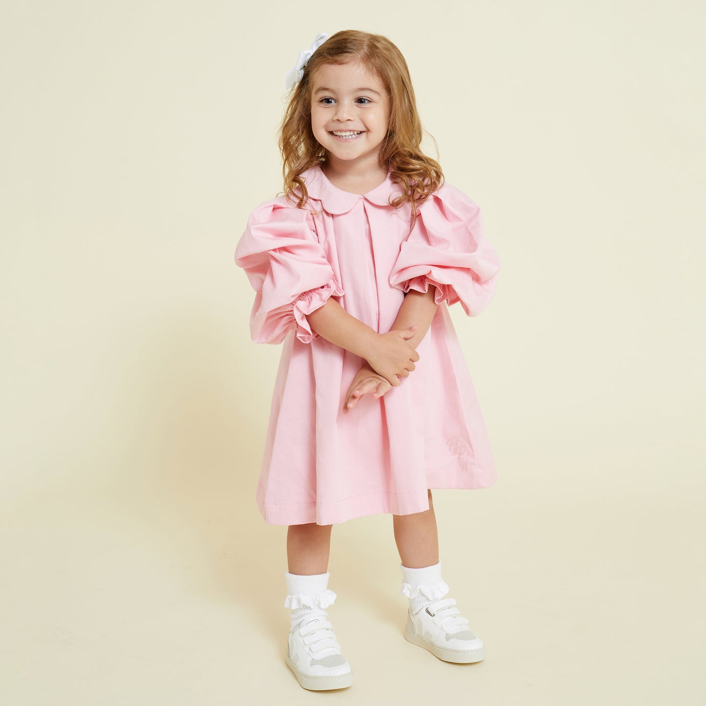 A girl aged 4 years old wearing a pink scallop collar smock dress with puff sleeves, paired with white socks and white shoes.