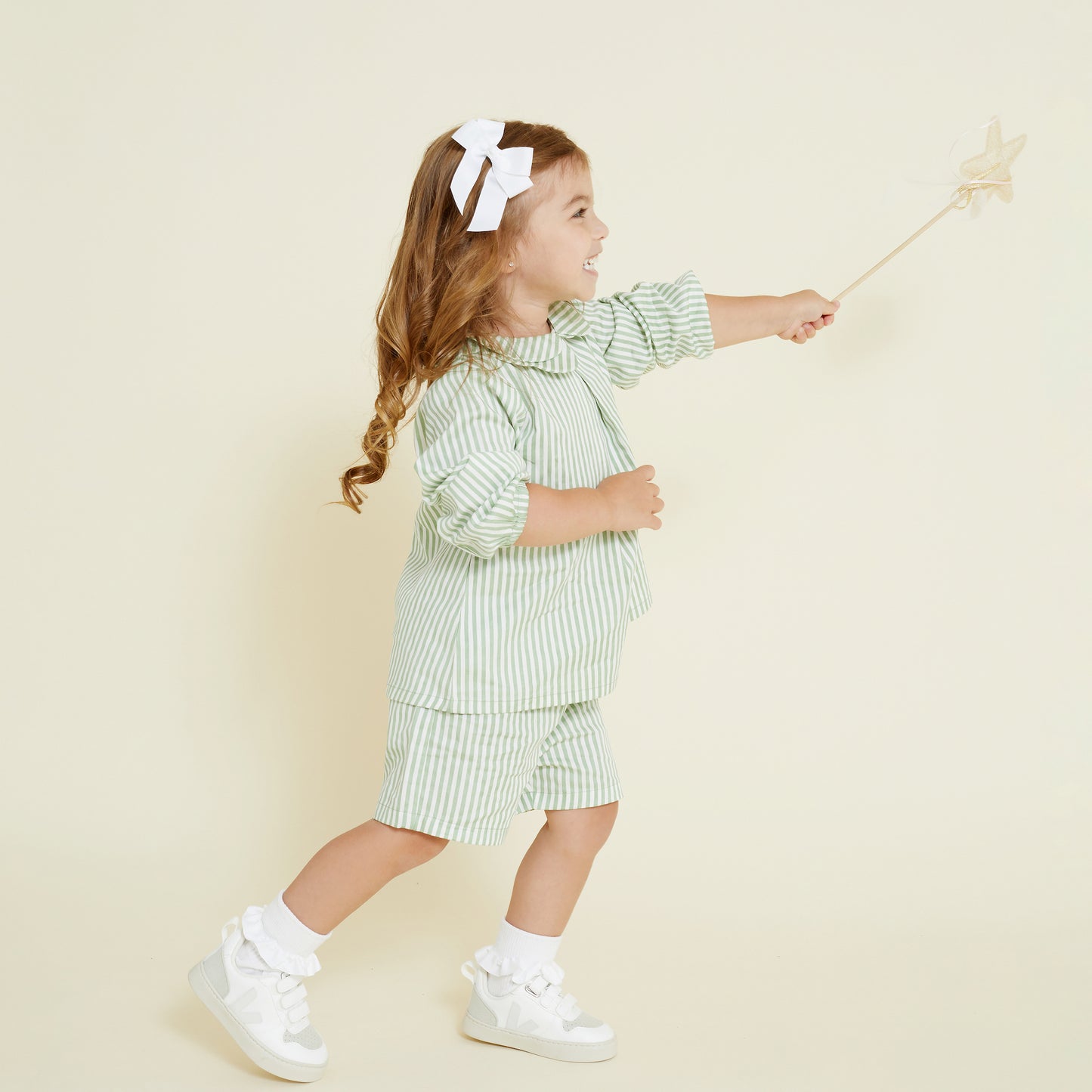 Young girl wears a matching set of collared shirt and casual short in a sage green and white stripe. She wears a mini hair bow in white.