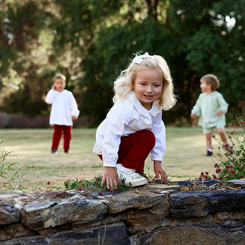 Little girl plays on a wall in the garden wearing white collared shirt and red pants