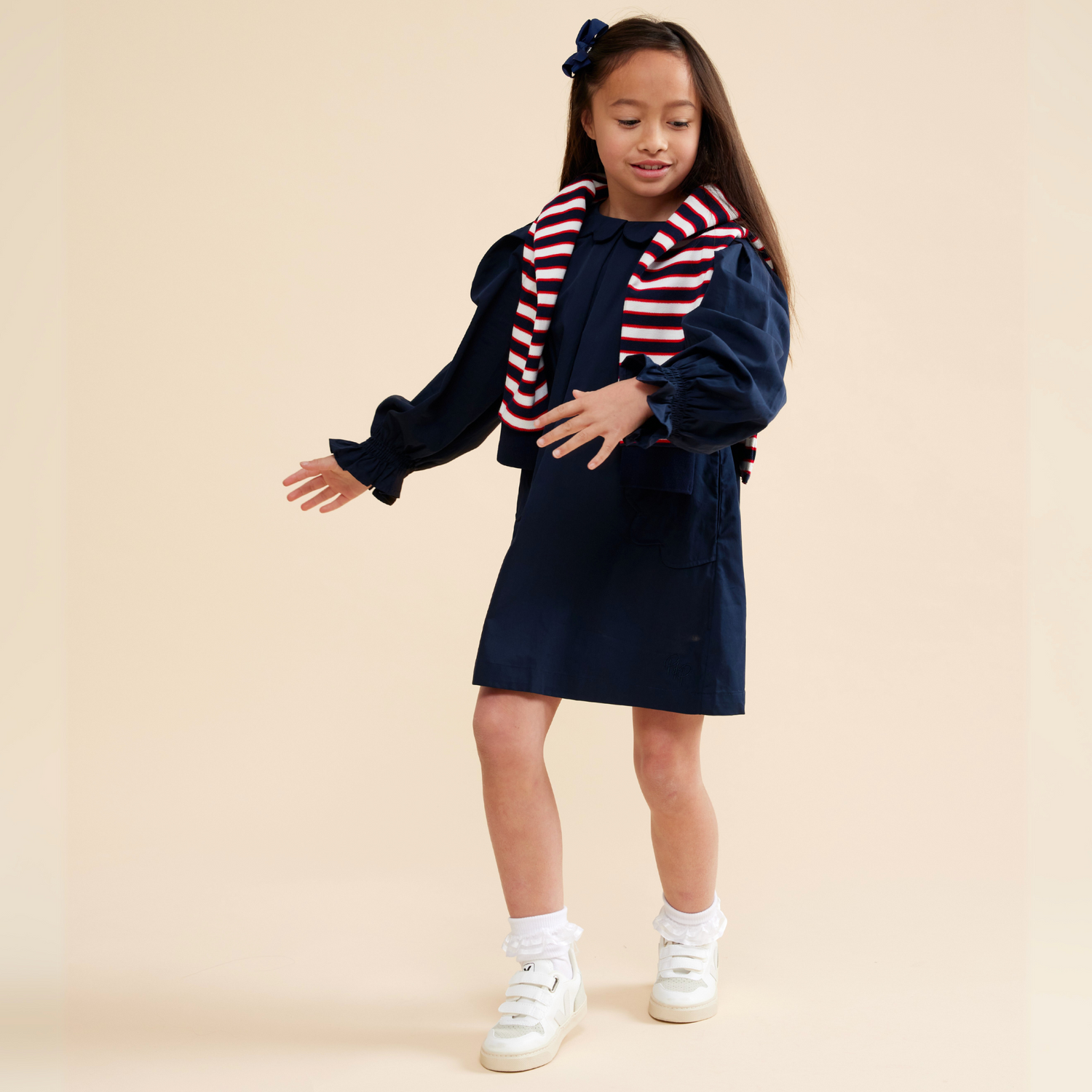 8 year old girl wearing navy scallop detail smock dress, paired with long sleeve polo t-shirt in navy, red and white stripe, She is doing a ballet twirl, wearing white socks and sneakers in front of a beige background. 