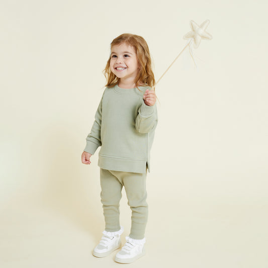 A 4 year old girl standing and holding a wand, wearing a sage green sweatshirt and matching pants