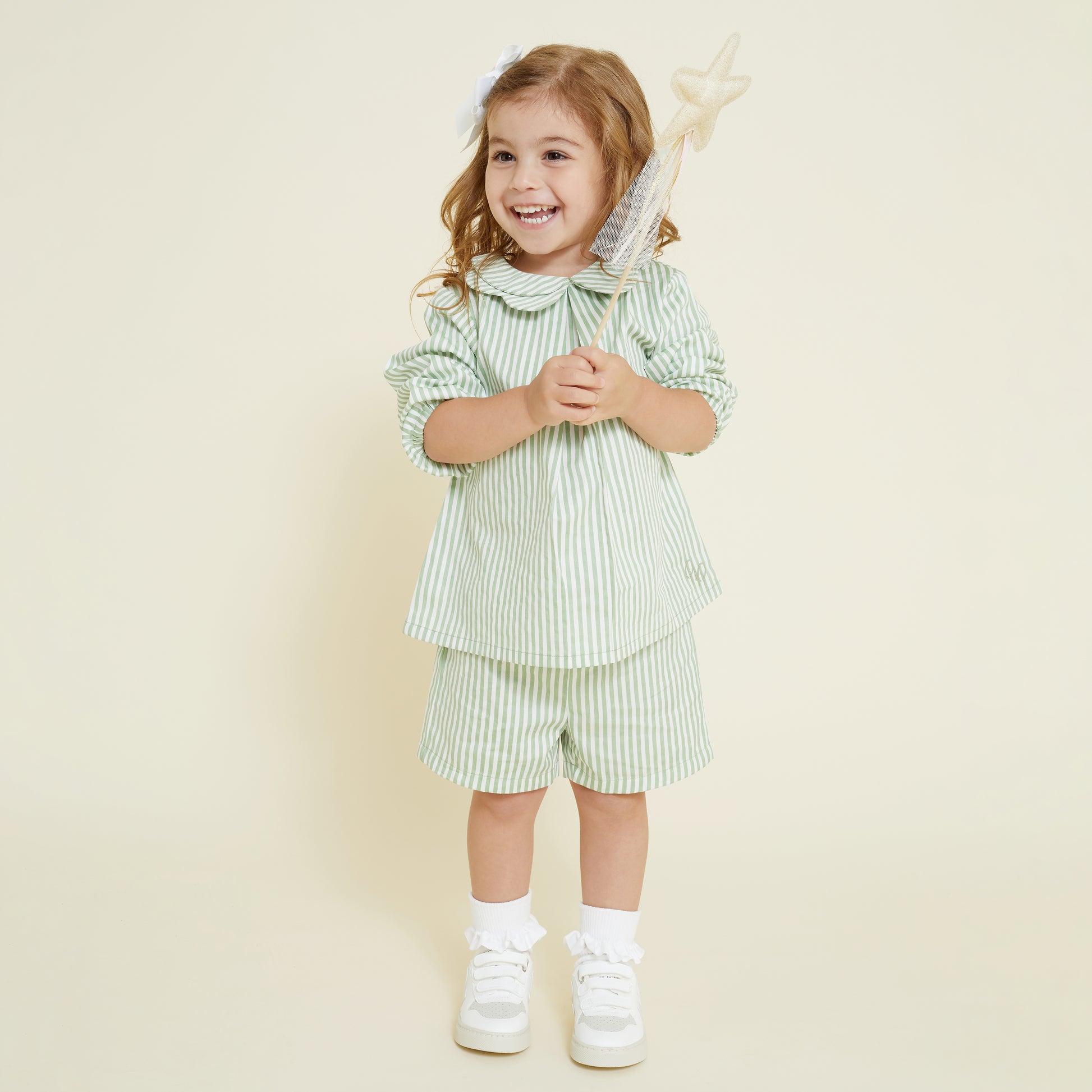 A 5 year old girl wearing a sage green and white striped collared shirt and matching pull on shorts, paired with white frill socks and white Veja sneakers. She holds a fairy wand in her hand. 