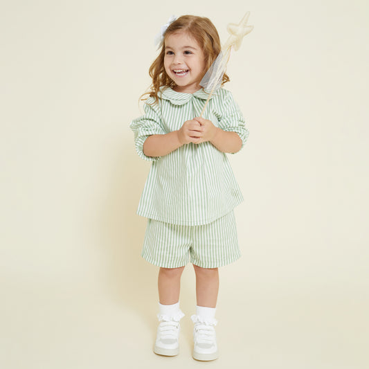 A 5 year old girl wearing a sage green and white striped collared shirt and matching pull on shorts, paired with white frill socks and white Veja sneakers. She holds a fairy wand in her hand. 