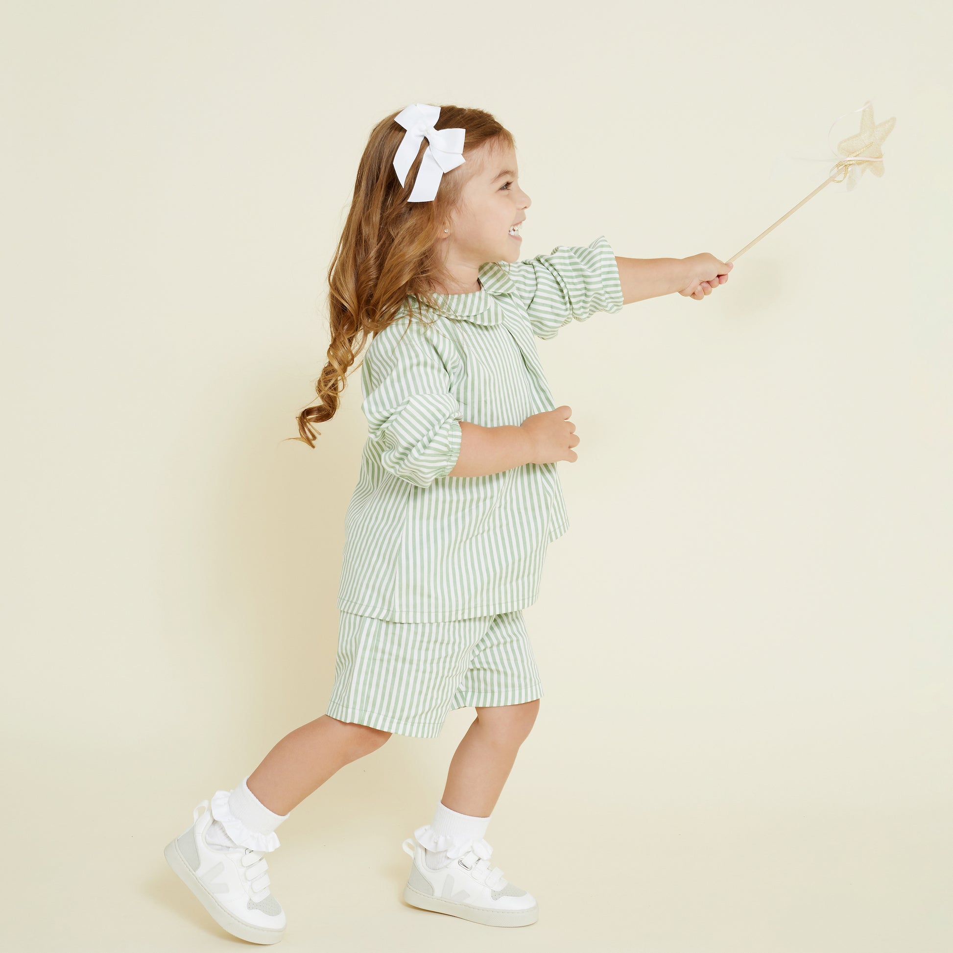 Young girl wears a matching set of collared shirt and casual short in a sage green and white stripe. She wears a mini hair bow in white. 
